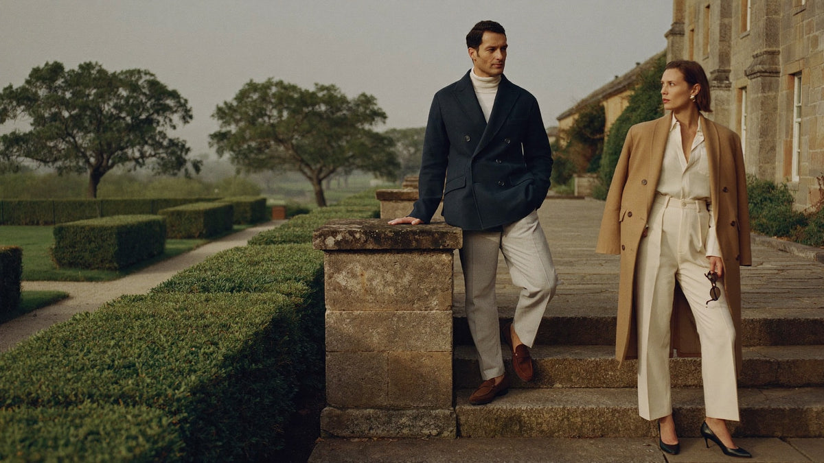 A man in a navy double-breasted blazer and a woman in a camel coat over cream trousers on the stone steps of a country house